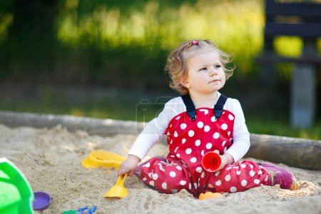 Cute toddler girl playing in sand on outdoor playground. Beautiful baby in red gum trousers having fun on sunny warm summer day. Child with colorful sand toys. Healthy active baby outdoors plays games