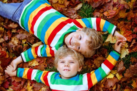 Téléchargez les photos : Deux petits jumeaux enfants garçons allongés dans des feuilles d'automne dans des vêtements de mode colorés. Heureux frères et sœurs qui s'amusent à Autumn Park par une journée chaude. Enfants en santé aux cheveux blonds au milieu du feuillage d'érable. - en image libre de droit