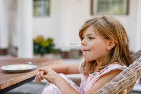 Photo for Cute preschool girl in nightgown sitting at breakfast table with family in summer garden. Happy child smiling and having fun - Royalty Free Image