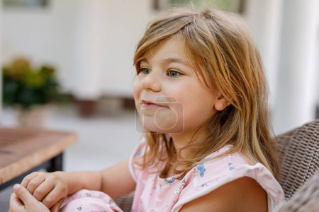 Téléchargez les photos : Jolie fille préscolaire en chemise de nuit assis à la table du petit déjeuner avec la famille dans le jardin d'été. Enfant heureux souriant et s'amusant - en image libre de droit