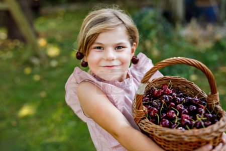 Foto de Hermosa chica en el jardín. Chica feliz con cerezas. Preschol niño con cesta llena de bayas maduras y divertidos pendientes de cereza - Imagen libre de derechos