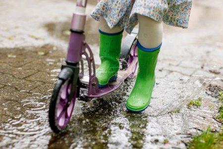 Téléchargez les photos : Fille sur un scooter par temps pluvieux. Enfant en chewing-gum dans les flaques d'eau après la pluie d'été. Du plaisir pour les enfants - en image libre de droit
