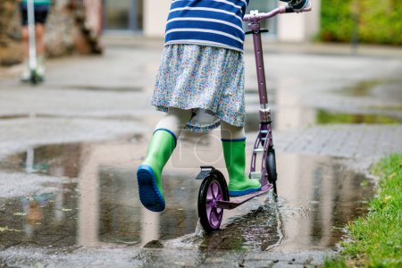 Photo for Girl on a scooter in rainy weather. Child in gum rainboots riding through puddles after summer rain. Fun for children - Royalty Free Image
