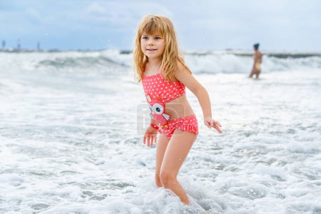 Téléchargez les photos : Enfant heureux, petite fille préscolaire en maillot de bain courant et sautant dans les vagues pendant les vacances d'été sur la plage tropicale exotique. Voyage en famille sur la côte de l'océan - en image libre de droit