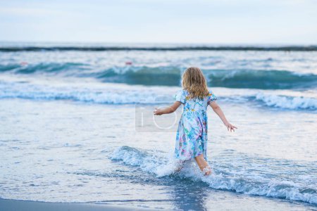 Téléchargez les photos : Enfant heureux, petite fille préscolaire en robe courant et sautant dans les vagues pendant les vacances d'été sur la plage tropicale exotique au coucher du soleil. Voyage en famille sur la côte de l'océan - en image libre de droit