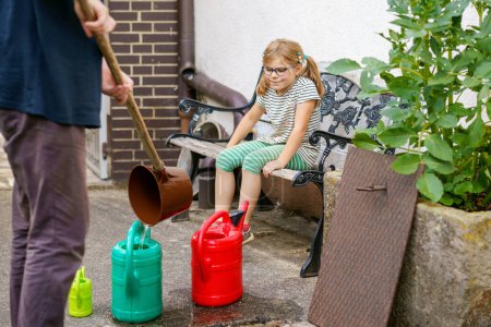 Téléchargez les photos : Little girl watering flowers in the garden. child waiting for father filling water in cans. Save water, drought,heat concept. Environment in europe or world. Rain water. - en image libre de droit