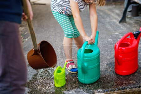 Foto de Little girl watering flowers in the garden. child waiting for father filling water in cans. Save water, drought,heat concept. Environment in europe or world. Rain water. - Imagen libre de derechos