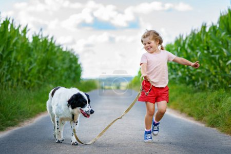 Téléchargez les photos : Enfant préscolaire Happy Girl avec chien courant sur la nature. Enfant souriant positif et meilleur ami animal de compagnie le jour ensoleillé de l'été. Animaux de famille et enfants, amour et amitié. - en image libre de droit