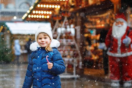 Photo for Little cute preschool girl with candy cane from a sweets stand on Christmas market. Happy child on traditional family market in Germany. Preschooler in colorful winter clothes during snowfall. - Royalty Free Image