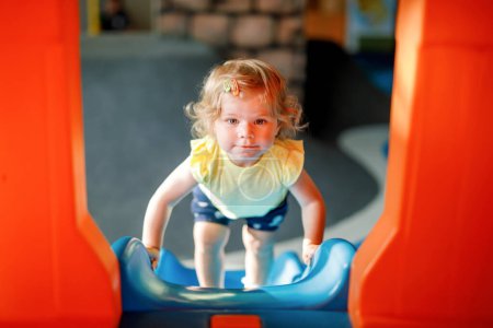 Photo for Happy blond little toddler girl having fun and sliding on indoor playground at daycare or nursery. Positive funny baby child smiling. Healthy girl climbing on slide - Royalty Free Image