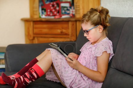 Photo for Positive little girl, holding tablet computer in her hands. Preschool child with eyeglasses playing games and learning with different apps - Royalty Free Image