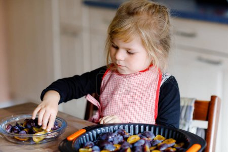 Photo for Cute little toddler girl baking plum pie at home. Happy smiling child helping and preparing plums for cake in domestic kitchen. Healthy homemade food - Royalty Free Image