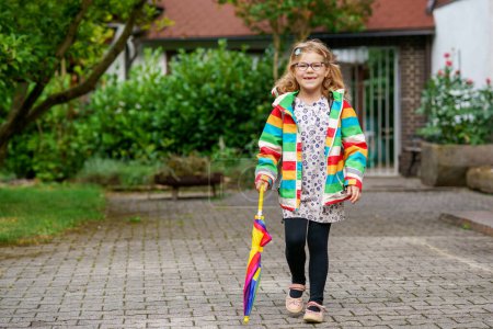 Photo for Little girl on way to elementary school or kindergarden. Preschool Child with colorful rainbow umbrella and waterproof jacket with school bag. Kid walking in autumn shower. Outdoor fun by any weather. - Royalty Free Image