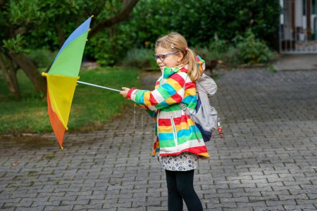Photo for Little girl on way to elementary school or kindergarden. Preschool Child with colorful rainbow umbrella and waterproof jacket with school bag. Kid walking in autumn shower. Outdoor fun by any weather. - Royalty Free Image