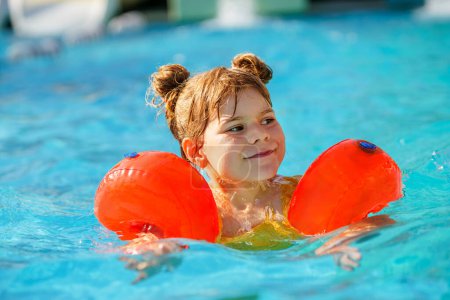 Photo for Little preschool girl with protective swimmies playing in outdoor swimming pool by sunset. Child learning to swim in outdoor pool, splashing with water, laughing and having fun. Family vacations - Royalty Free Image