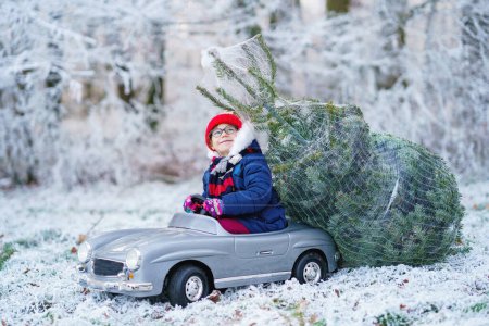 Foto de Feliz niña sonriente conduciendo coche de juguete con árbol de Navidad. Divertido niño preescolar en ropa de invierno que trae el árbol de Navidad tallado de bosque nevado. Familia, tradición, vacaciones - Imagen libre de derechos