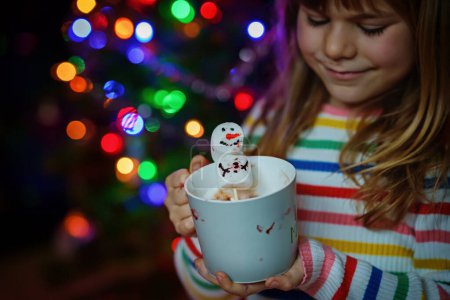 Foto de Niña sosteniendo taza con chocolate caliente con malvaviscos como muñeco de nieve. Niño sentado cerca del árbol de Navidad decorado con luces - Imagen libre de derechos