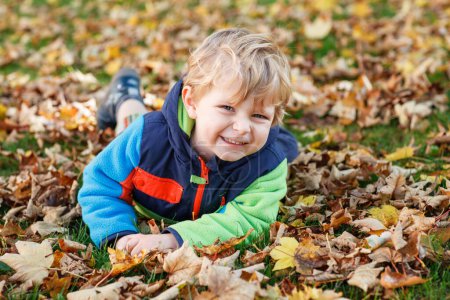 Téléchargez les photos : Joli petit garçon jouant avec des feuilles d'érable à l'extérieur. Joyeux enfant marchant dans le parc d'automne. Bébé garçon enfant porte une veste à la mode. portrait de garçon blond souriant. Mode d'automne. Enfant élégant dehors - en image libre de droit