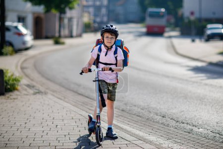 Foto de Niño de escuela activa con gafas en casco de seguridad montando con su scooter en la ciudad con mochila en un día soleado. Feliz niño en bicicleta de camino a la escuela. Manera segura para los niños al aire libre a la escuela - Imagen libre de derechos
