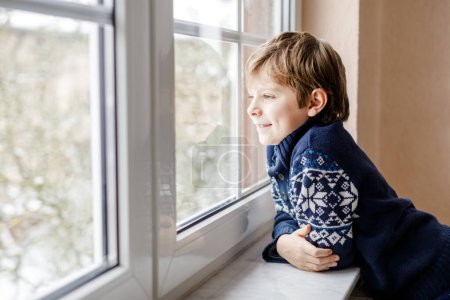 Foto de Niño adorable feliz sentado cerca de la ventana y mirando hacia fuera en la nieve en el día de Navidad o la mañana. Sonriente niño sano fascinado observando nevadas y grandes copos de nieve. - Imagen libre de derechos