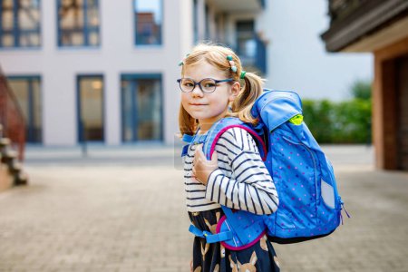 Photo for Cute little girl on her first day going to school. Healthy beautiful child walking to nursery preschool and kindergarten. Happy child with eyeglasses with backpack on the city street, outdoors - Royalty Free Image