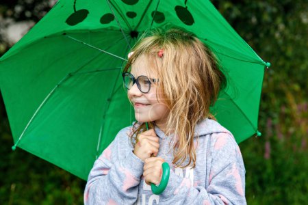 Foto de Niña preescolar caminando durante la lluvia con un gran paraguas verde en el día lluvioso. Feliz niño positivo. Actividad de los niños en el mal tiempo - Imagen libre de derechos