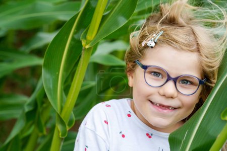 Photo for Portrait of happy little girl hiding in a field among green corn stalks. Positive preschool child having fun outdoors, summer activity - Royalty Free Image