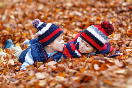 Foto de Dos niñitos gemelos acostados en otoño se van con ropa colorida. Felices hermanos niños divirtiéndose en el bosque de otoño o parque en el día de otoño. Con sombreros y bufandas de moda casual. Amigos jugando juntos - Imagen libre de derechos