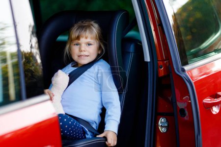 Photo for Adorable toddler girl sitting in car seat, holding plush soft toy and looking out of the window on nature and traffic. Little kid traveling by car. Child safety on the road. Family trip and vacations. - Royalty Free Image