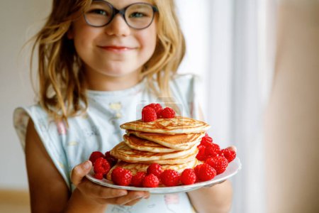 Photo for Little happy preschool girl with a large stack of pancakes and raspberries for breakfast. Positive child eating healthy homemade food in the morning - Royalty Free Image