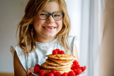 Téléchargez les photos : Petite fille préscolaire heureuse avec une grande pile de crêpes et de framboises pour le petit déjeuner. Enfant positif mangeant des aliments maison sains le matin - en image libre de droit