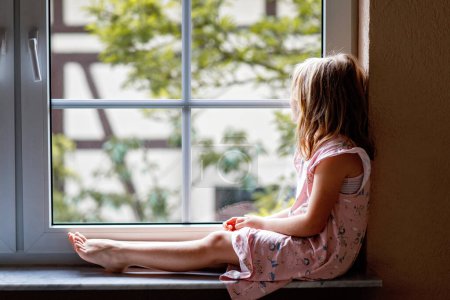 Téléchargez les photos : Adorable petite fille assise près de la fenêtre. préscolaire enfant regardant dehors et rêvant - en image libre de droit