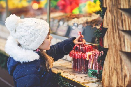 Téléchargez les photos : Petite fille mignonne préscolaire achetant canne à bonbons à partir d'un stand de bonbons sur le marché de Noël. Enfant heureux sur le marché familial traditionnel en Allemagne. Préscolaire en vêtements d'hiver colorés. - en image libre de droit