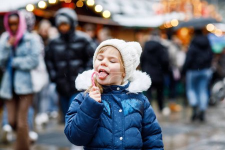 Téléchargez les photos : Petite fille mignonne préscolaire avec canne à bonbons d'un stand de bonbons sur le marché de Noël. Enfant heureux sur le marché familial traditionnel en Allemagne. Préscolaire en vêtements d'hiver colorés pendant les chutes de neige. - en image libre de droit