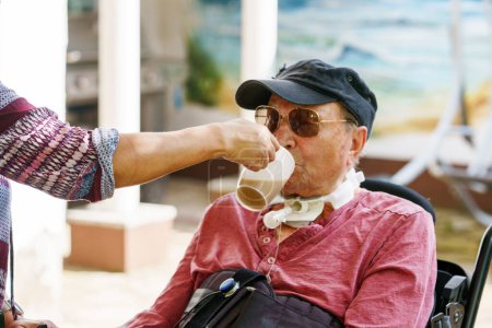 Téléchargez les photos : Un aîné de 85 ans en fauteuil roulant électrique est assis dans un parc. Infirmière soutenant l'homme avec boire du café, tenant tasse - en image libre de droit