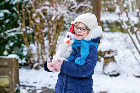 Foto de Cute little preschool girl with glasses making mini snowman. Adorable healthy happy child playing and having fun with snow, outdoors on cold day. Active leisure with children in winter. - Imagen libre de derechos