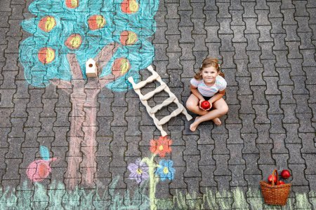 Téléchargez les photos : Jolie petite fille cueillette des pommes de pommier peint avec des craies colorées sur l'asphalte. Mignon enfant d'âge préscolaire avec s'amuser avec une image à la craie. Loisirs créatifs pour les enfants, dessin et peinture - en image libre de droit
