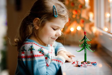 Photo for Litte toddler girl sitting by window and decorating small glass Christmas tree with tiny xmas toys. Happy healthy child celebrate family traditional holiday. Adorable baby - Royalty Free Image