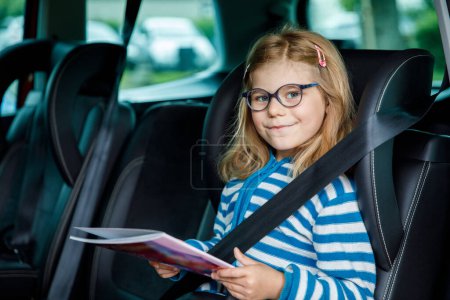 Téléchargez les photos : Petite fille d'âge préscolaire assise dans son siège auto. Happy Child avec des lunettes de vue Lire un livre, sourire sur le chemin des vacances en famille pendant les embouteillages - en image libre de droit