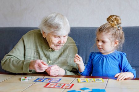 Foto de Hermosa niña y abuela jugando juntos fotos lotería juego de cartas de mesa en casa. Lindo niño y mujer mayor divirtiéndose juntos. Familia feliz en el interior
. - Imagen libre de derechos
