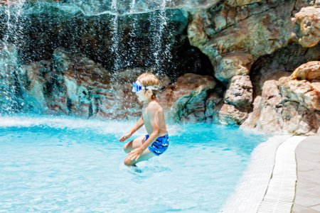 Téléchargez les photos : Joyeux petit garçon sautant dans la piscine et s'amusant en vacances en famille dans une station hôtelière. Enfant sain jouant dans l'eau - en image libre de droit