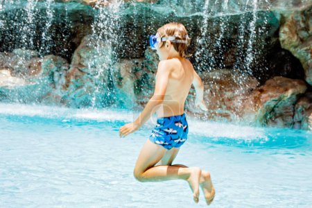 Foto de Feliz niño saltando en la piscina y divirtiéndose en vacaciones familiares en un complejo hotelero. Niño sano jugando en el agua - Imagen libre de derechos