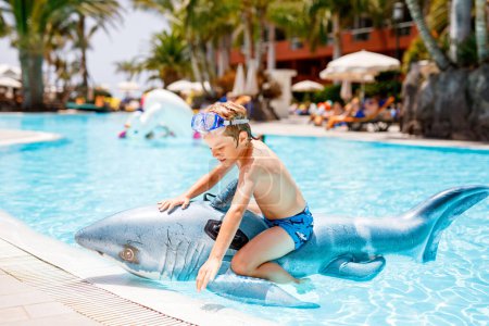 Téléchargez les photos : Joyeux petit garçon sautant dans la piscine et s'amusant en vacances en famille dans une station hôtelière. Enfant sain jouant dans l'eau avec le jouet gonflable de requin - en image libre de droit