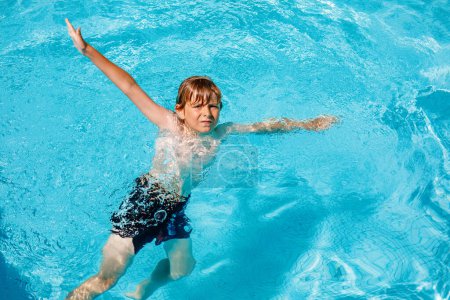 Téléchargez les photos : Portrait de petit garçon heureux dans la piscine et s'amuser en vacances en famille dans une station hôtelière. Enfants en bonne santé jouant dans l'eau, nageant et éclaboussant - en image libre de droit