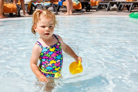 Téléchargez les photos : Jolie petite fille heureuse tout-petit dans la piscine et s'amuser en vacances en famille dans une station hôtelière. Enfant en bonne santé jouant dans l'eau. Bébé en maillot de bain coloré - en image libre de droit