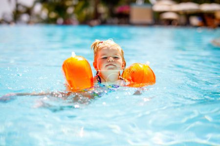 Photo for Little toddler girl with protective swimmies playing in outdoor swimming pool by sunset. Baby Child learning to swim in outdoor pool, splashing with water, laughing and having fun. Family vacations - Royalty Free Image