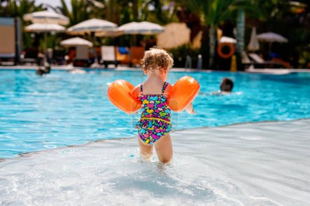 Téléchargez les photos : Petite fille tout-petit avec des nageurs protecteurs jouant dans la piscine extérieure au coucher du soleil. Bébé Enfant apprenant à nager dans la piscine extérieure, éclaboussant d'eau, riant et s'amusant. Vacances en famille - en image libre de droit