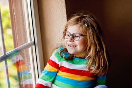 Foto de Adorable niña preescolar con anteojos sentados junto a la ventana. Niño reflexivo mirando hacia fuera. Chico solitario - Imagen libre de derechos