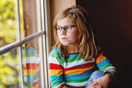 Téléchargez les photos : Adorable petite fille d'âge préscolaire avec des lunettes assis près de la fenêtre. Enfant réfléchi regardant dehors. Enfant solitaire - en image libre de droit