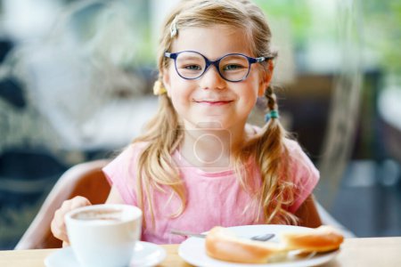 Foto de Little smiling girl have a breakfast in a cafe. Preschool child with glasses drinking hot chocolate and eating bakery pastry croissant or cake. Happy children, healthy food and meal - Imagen libre de derechos